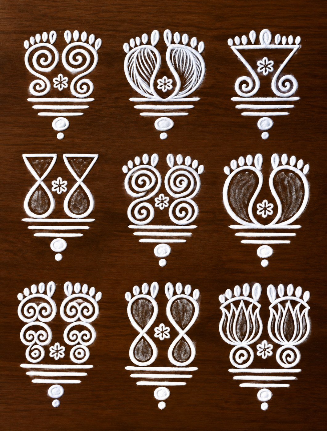 10 beautiful rangoli design ideas for entrances, stairs and floors Nine traditional white rangoli motifs arranged in three rows on a dark brown background, featuring paired footprints, loops, dots and curved ornaments.