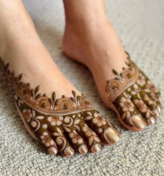 Feet with brown mehndi featuring rounded floral motifs, curved bands and decorated toes